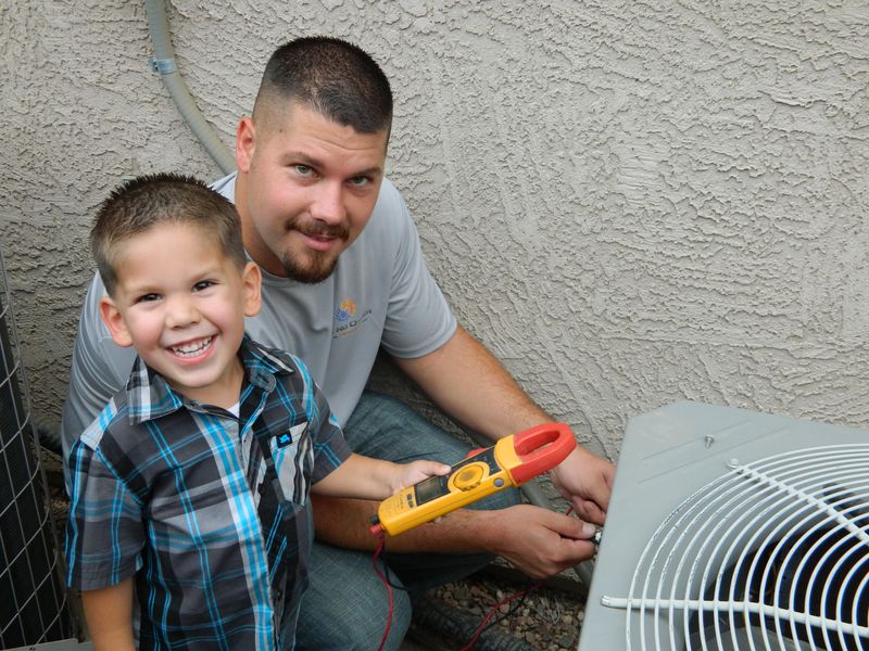 Kris kneeling at an AC condenser with his young son, who is smiling and holding a clamp meter