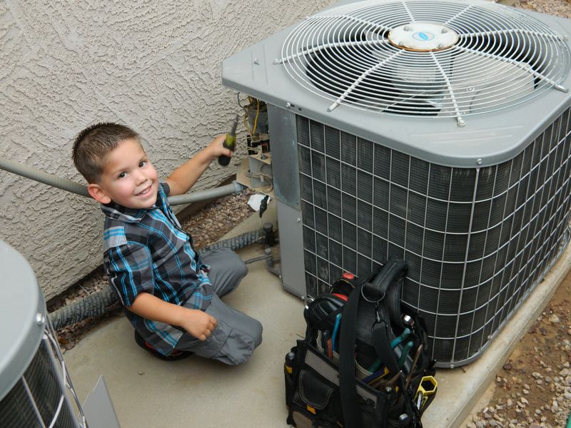 Kris&rsquo;s young son kneeling at an AC condenser with a screwdriver, smiling, with a tool bag beside him
